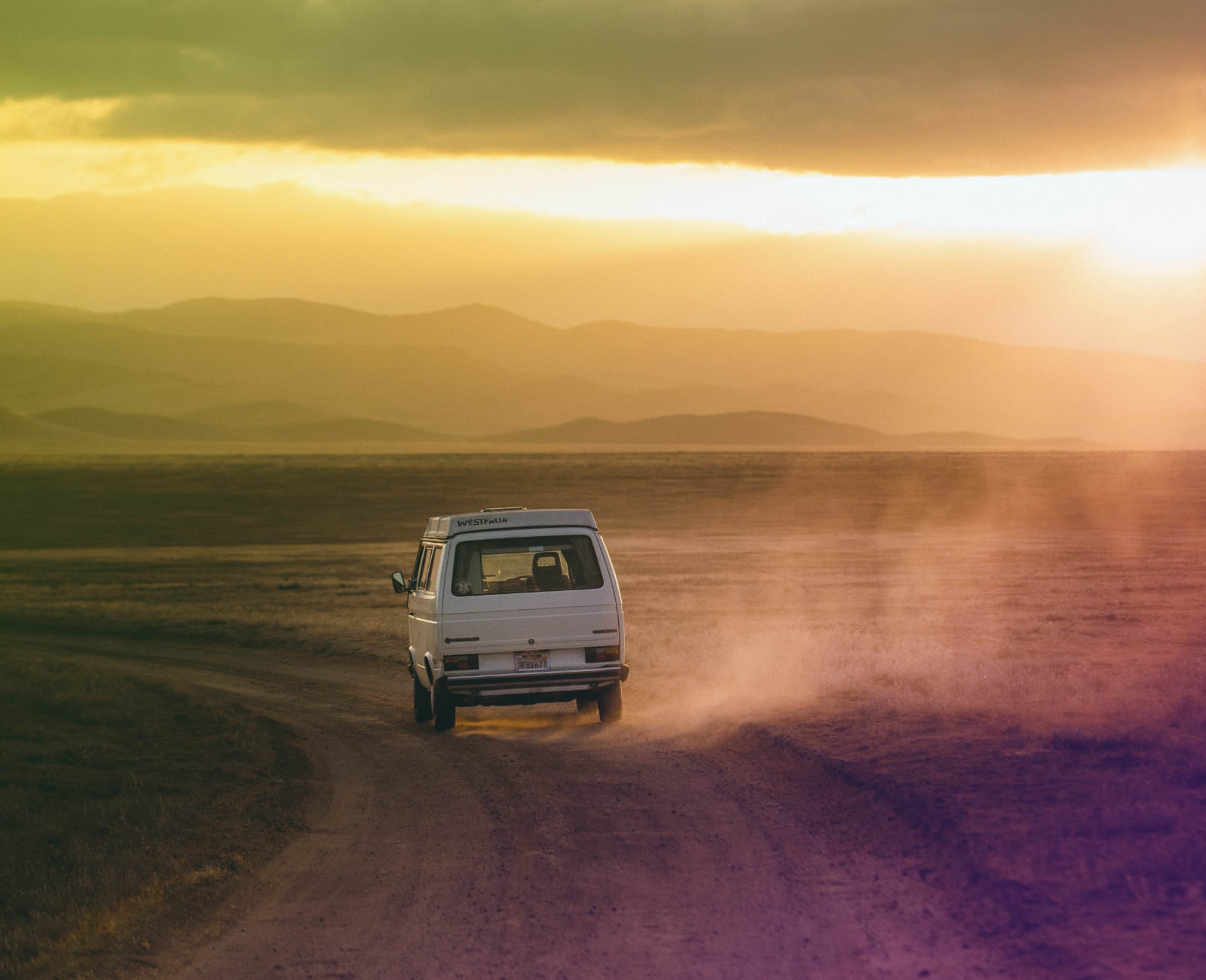 Bild von einem alten Camping-Bus, der durch eine Wüstenlandschaft fährt und dabei Staub aufwirbelt.